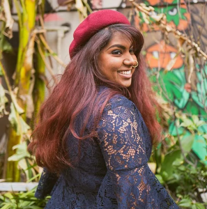 Nisha has a red beret atop her head and wears a big smile as she glances over her shoulder at the camera. She has long reddish brown hair, dark eyes and a nosering. She wears a royal blue/purple lace top and stands in front of a mural and green leafy backdrop.